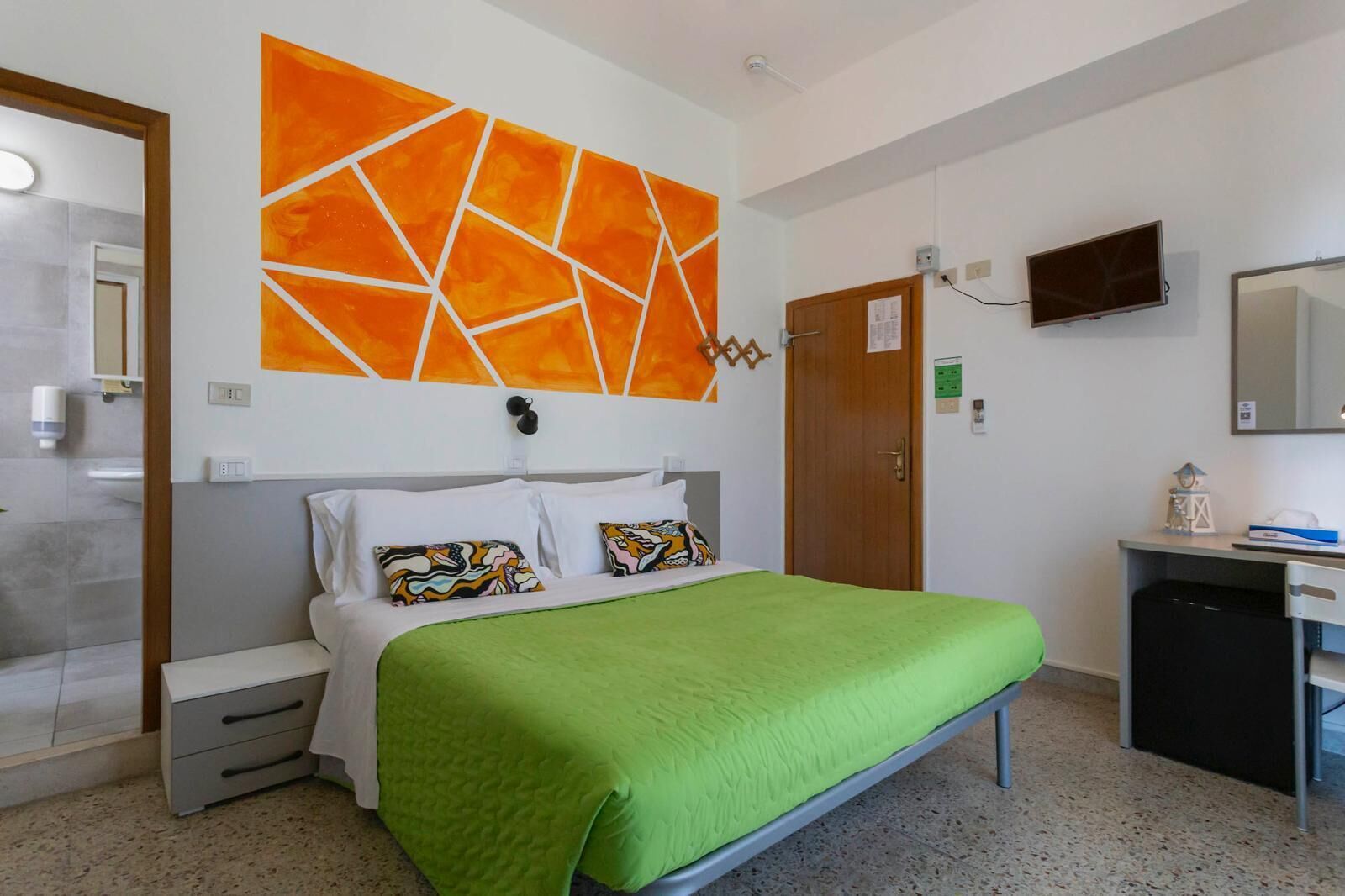 A hotel room with a bed, a desk, a TV, and a large orange abstract painting on the wall.