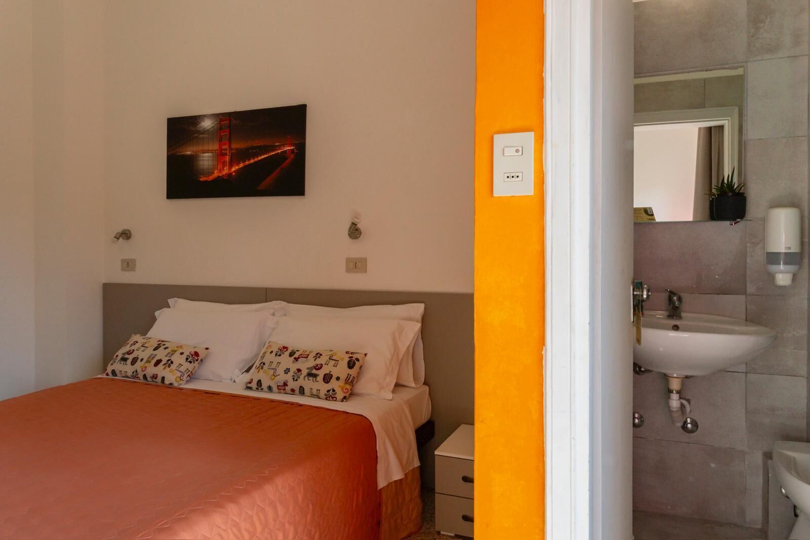 Un dormitorio con cama, almohadas, una mesita de noche, un cuadro, y un baño con lavamanos y espejo.