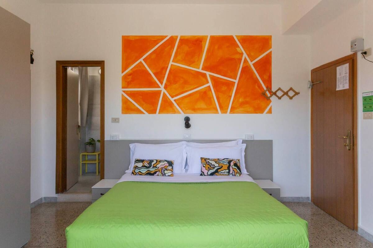 A hotel room with a bed, a green bedspread, two pillows with colorful patterns, and a large orange abstract painting on the wall.