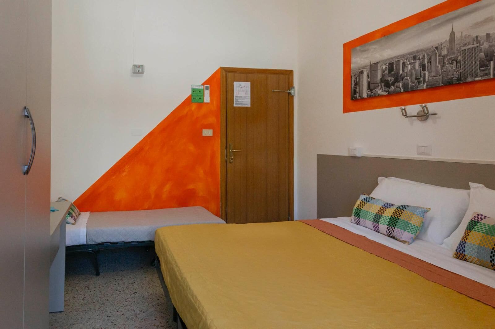 Una stanza con due letti, uno con copriletto giallo e l'altro con copriletto bianco. Si vede una porta di legno e un quadro appeso al muro.