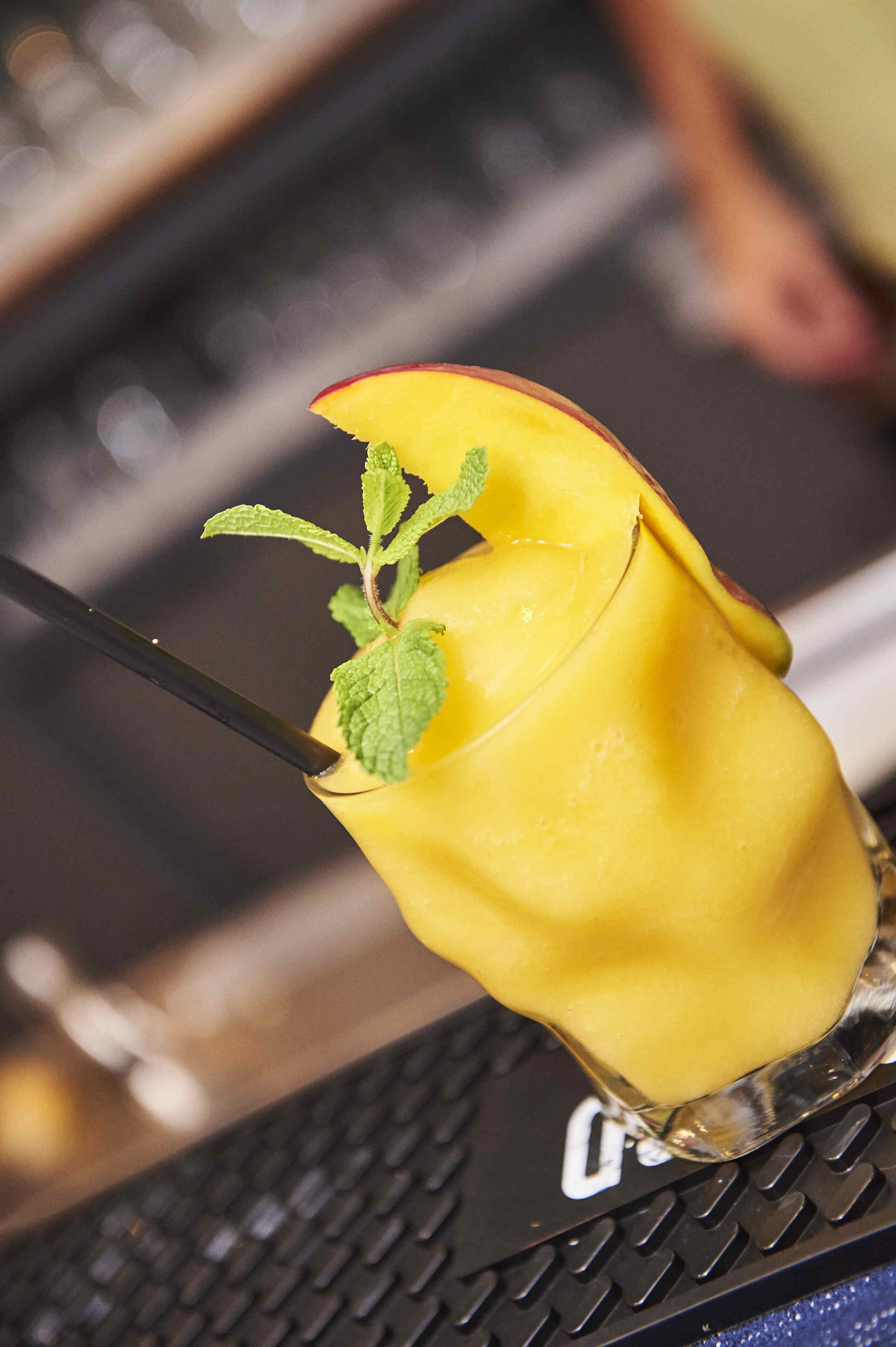 Un verre contenant une boisson jaune, agrémenté d’une feuille de menthe et d’une tranche de fruit, posé sur une surface noire texturée.