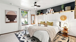 Ein Schlafzimmer mit einem Bett, einem Deckenventilator, einem Fenster, einem Nachttisch und einem an der Wand befestigten Regal mit Dekoration.