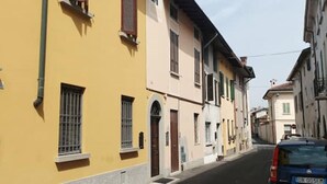 A narrow street with yellow and white buildings, a blue car parked on the right, and a wooden door on the left.