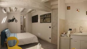 A compact bedroom with a bed, a chair, a small bathroom with a sink, and a motorcycle decal on the wall.