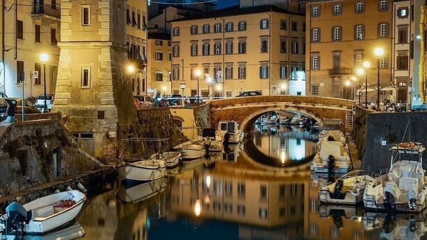 A nighttime view of a canal with a bridge, boats, and buildings reflecting on the water.