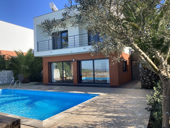 A modern two-story house with a swimming pool, large glass doors, and a balcony.