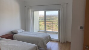Een slaapkamer met twee bedden, uitzicht op een groen landschap door een groot raam, en een houten vloer.