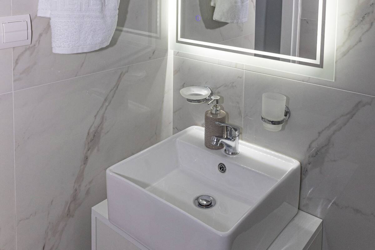 A bathroom with a white sink, a mirror with built-in lighting, and marble walls.