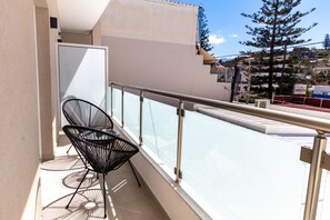 Ein Balkon mit zwei schwarzen Stühlen, einer Glasreling und Blick auf einen Baum sowie Gebäude im Hintergrund.