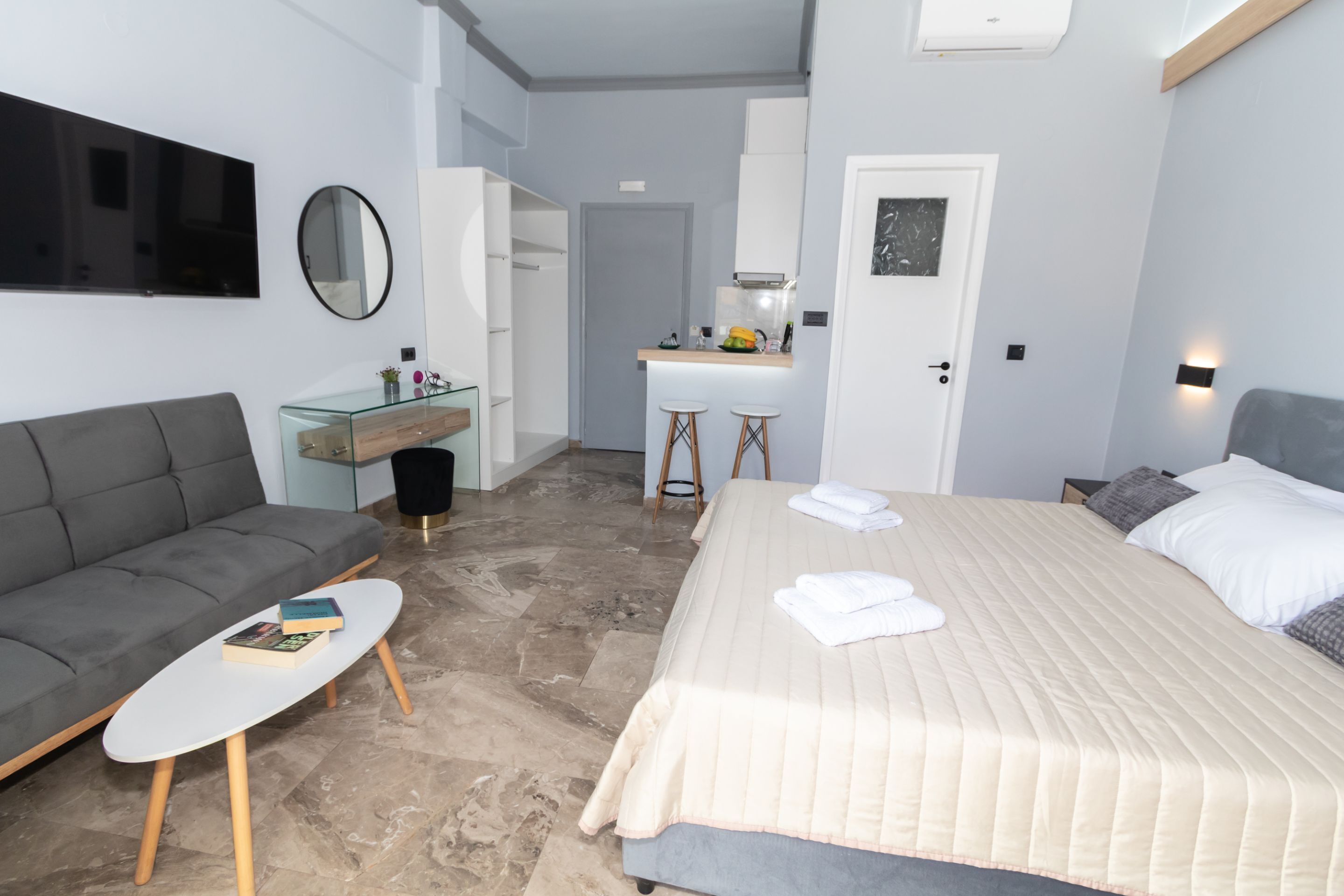 Une chambre d’hôtel moderne avec un canapé gris, une petite table blanche, un lit recouvert d’une courtepointe beige, une table basse en verre, un coin cuisine avec des tabourets et une porte blanche.