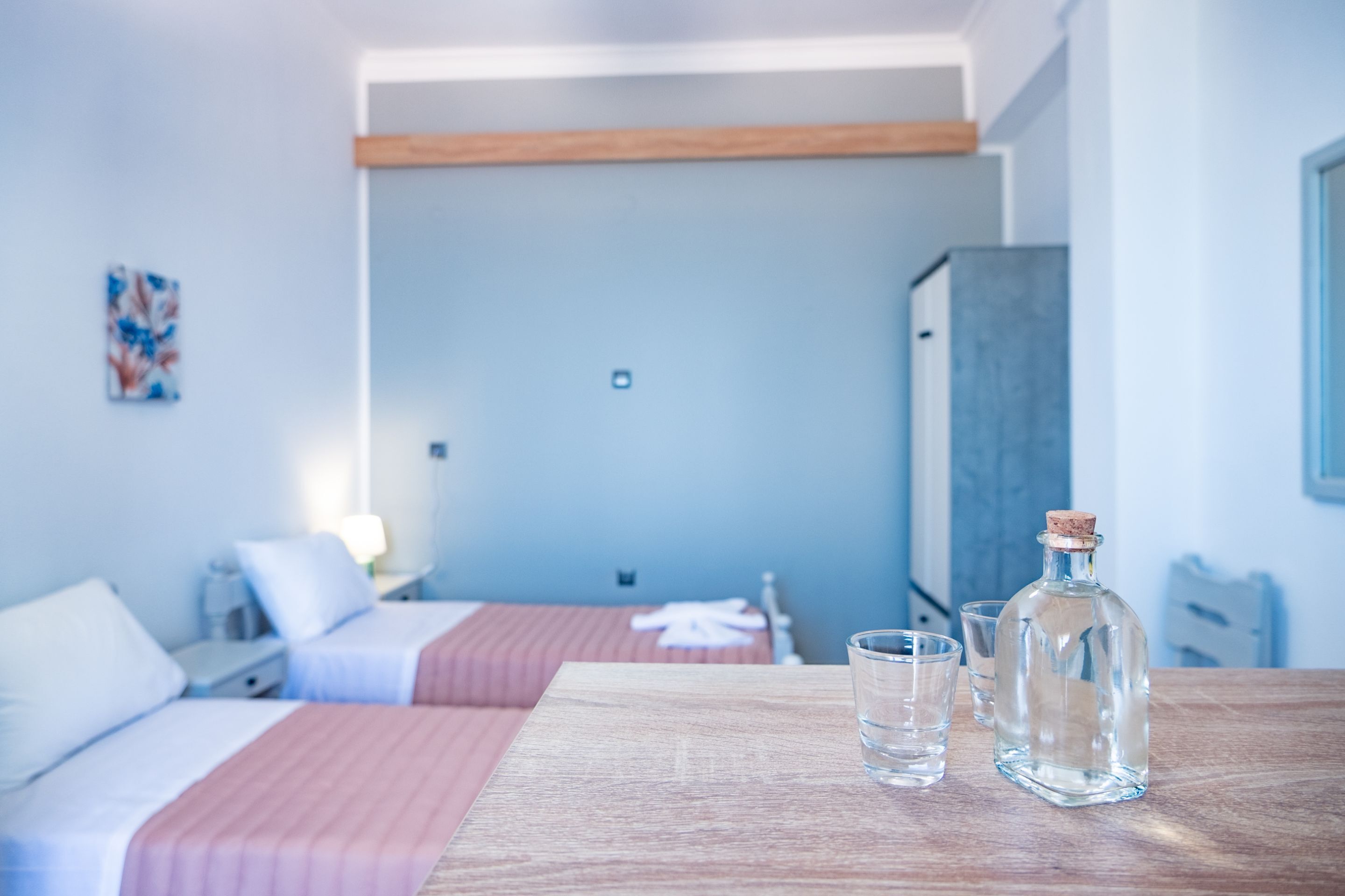 A hotel room with two beds, a wooden table with a bottle and glasses, and a painting on the wall.