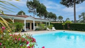 A poolside area with a covered patio, lounge chairs, and a view of a house with a tiled roof. - Swimming pool villa for 8 in Lacanau (Lacanau)
