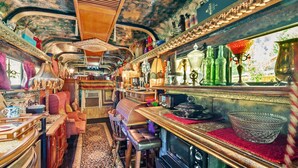 A vintage train carriage interior with wooden furniture, a dining area, and a kitchen with a stove.