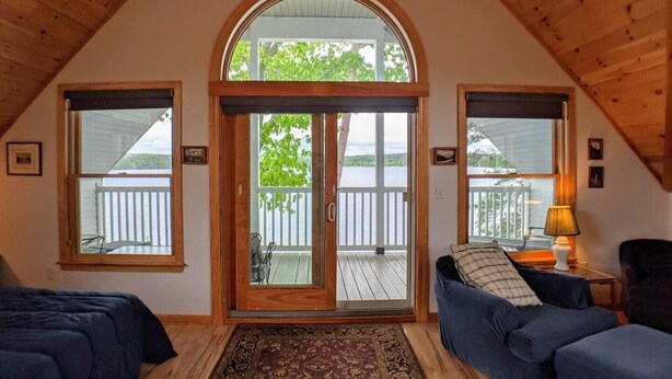 Une pièce chaleureuse avec un plafond voûté, une grande porte en arc donnant sur un balcon et une vue sur un lac. On y trouve deux canapés bleus et une petite table avec une lampe.