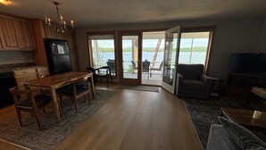 Un vaste espace de vie comprenant une table et des chaises en bois, un canapé et un téléviseur. Une porte-fenêtre coulissante donne accès à un balcon avec vue sur un lac.