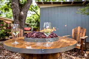 Deux verres à vin sur une table en bois ornée d’un anneau métallique, un foyer avec des bûches, et une chaise en bois dans un jardin.
