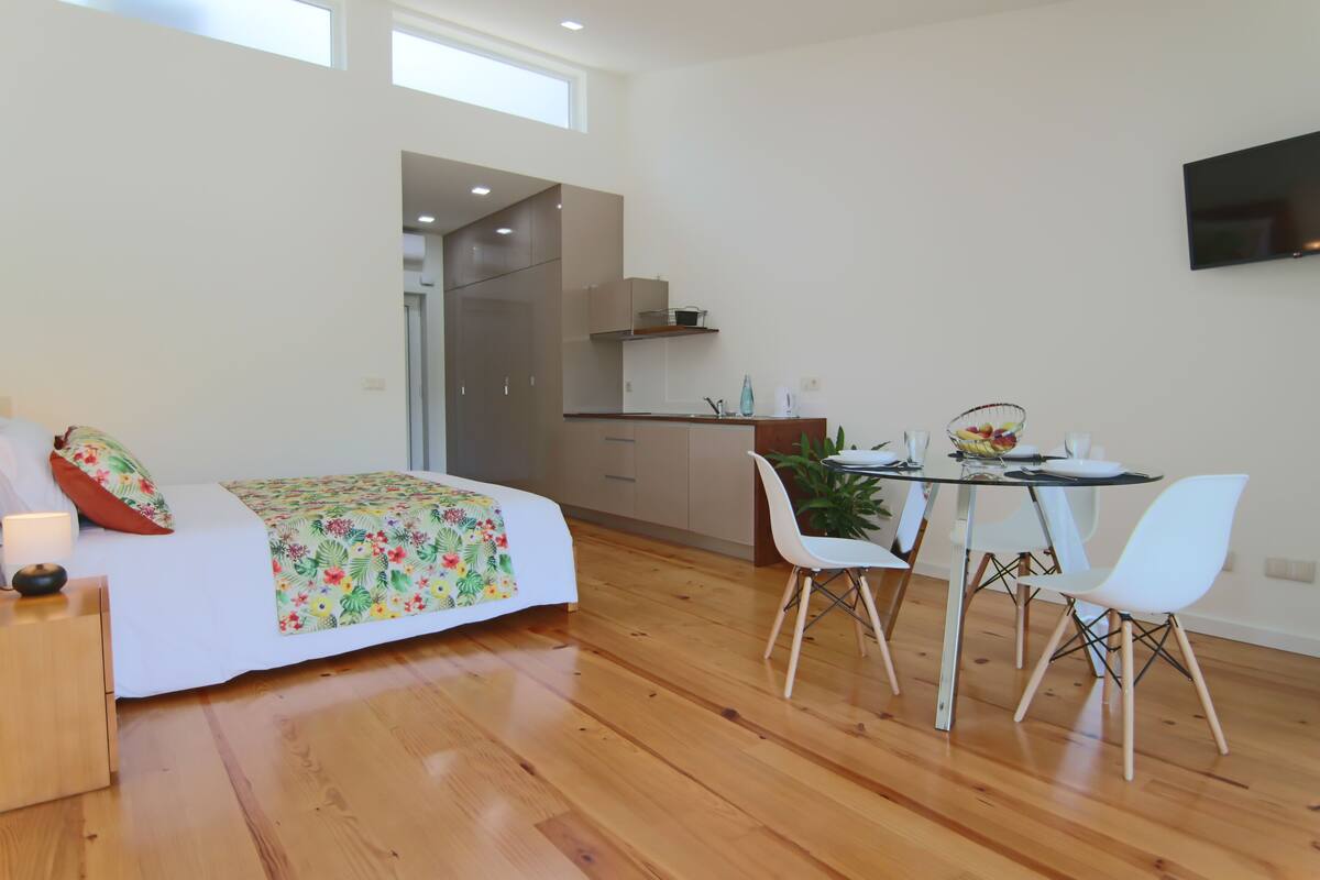 A modern room with a bed, a dining table with chairs, a TV mounted on the wall, and a kitchen area.
