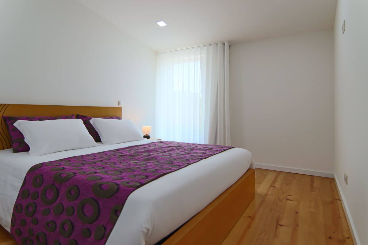 A neatly made bed with a purple and white patterned bedspread, a wooden headboard, and a nightstand with a lamp.