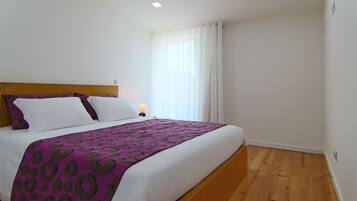 Een net opgemaakt bed met een paars-witte spreideken, een houten hoofdbord en een nachtkastje met een lamp.