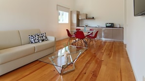 A modern living room with a glass coffee table, a sofa, and red chairs. There is a kitchen area with cabinets and a microwave.