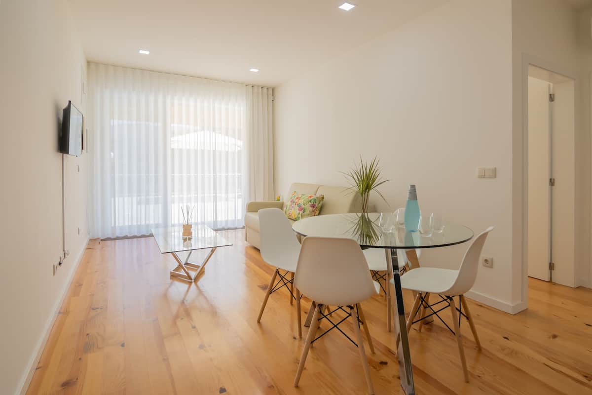 A modern dining area with a glass table, white chairs, and a sofa with a floral pillow.