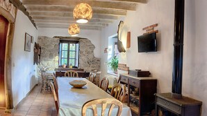 Un coin repas rustique avec une longue table, des chaises en bois, un mur en pierre, une cheminée et un téléviseur fixé au mur.