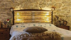 Une chambre avec un mur en pierre, une tête de lit en bois agrémentée de panneaux lumineux, une couvre-lit à motifs floraux et une table de chevet sur laquelle est posé un vase de fleurs.