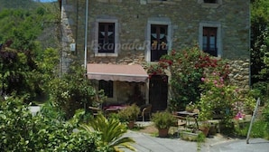 Un bâtiment en pierre avec un toit en tuiles, un petit porche avec une table et des chaises, et un jardin agrémenté de diverses plantes et fleurs.