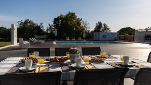 Ein Essbereich im Freien mit einem Pool im Hintergrund, ein Tisch ist für das Frühstück gedeckt, mit Tellern, Gläsern und einer Blumenvase.