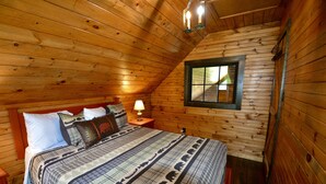 A cozy wooden cabin bedroom with a large bed, a nightstand, a lamp, and a window.