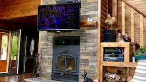 A log cabin interior with a stone fireplace, a mounted television, and a wooden ceiling.