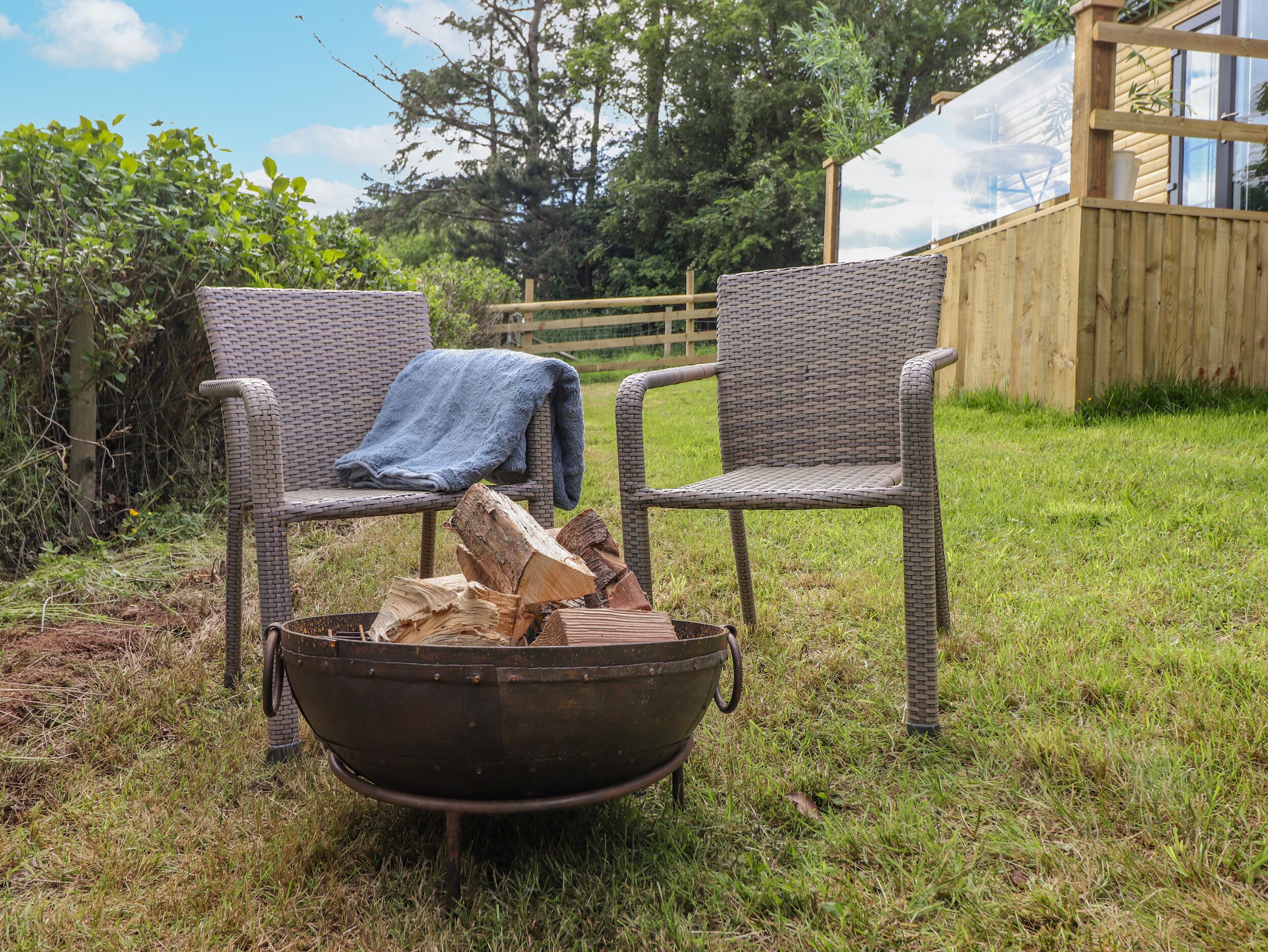 Two wicker chairs with a blanket on one, a metal fire pit with logs, and a wooden fence in a grassy outdoor area.