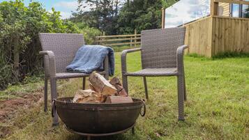 Two wicker chairs with a blanket on one, a metal fire pit with logs, and a wooden fence in a grassy outdoor area.