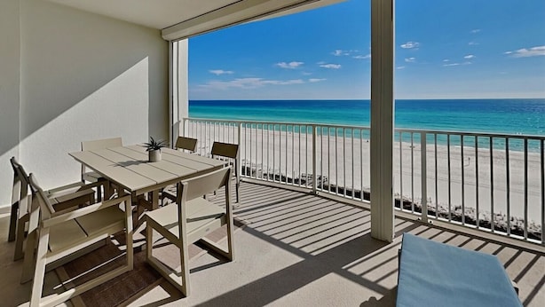 A balcony with a table and chairs, a view of the beach, and a clear sky.