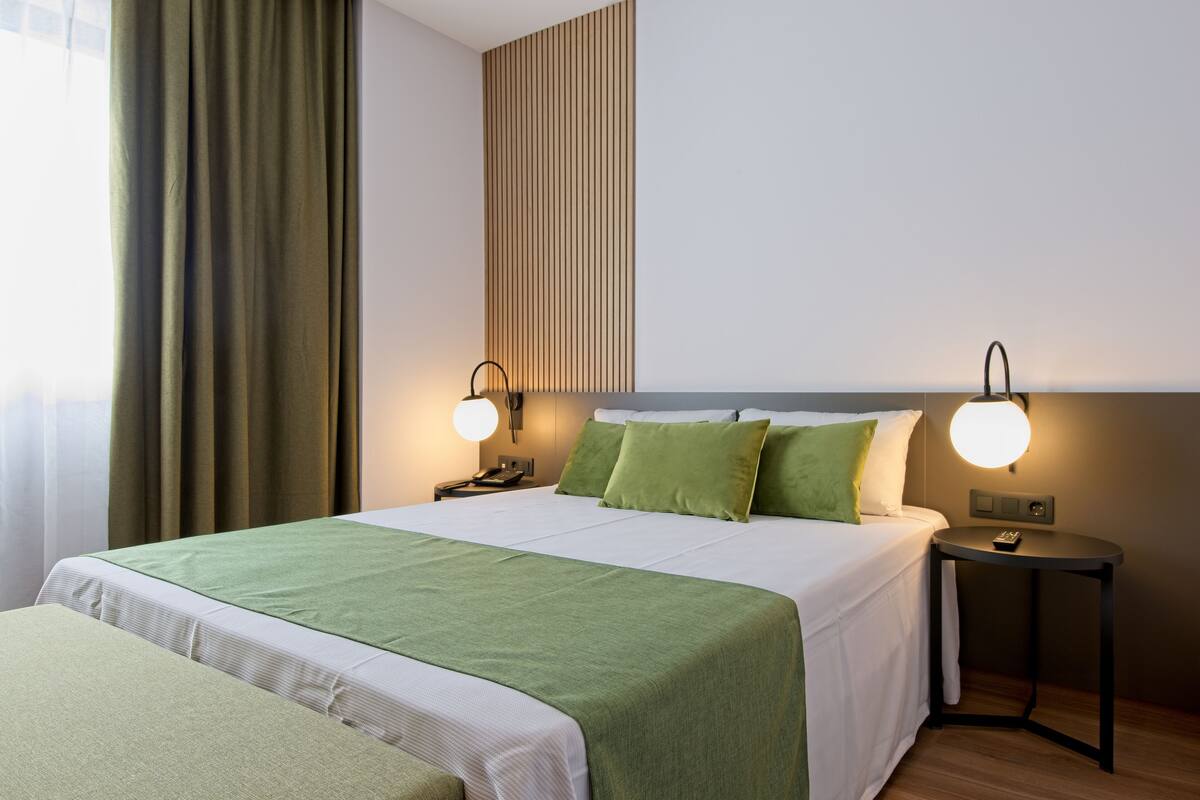 A hotel room with a neatly made bed, two bedside tables with lamps, a window with curtains, and a wall with vertical lines.