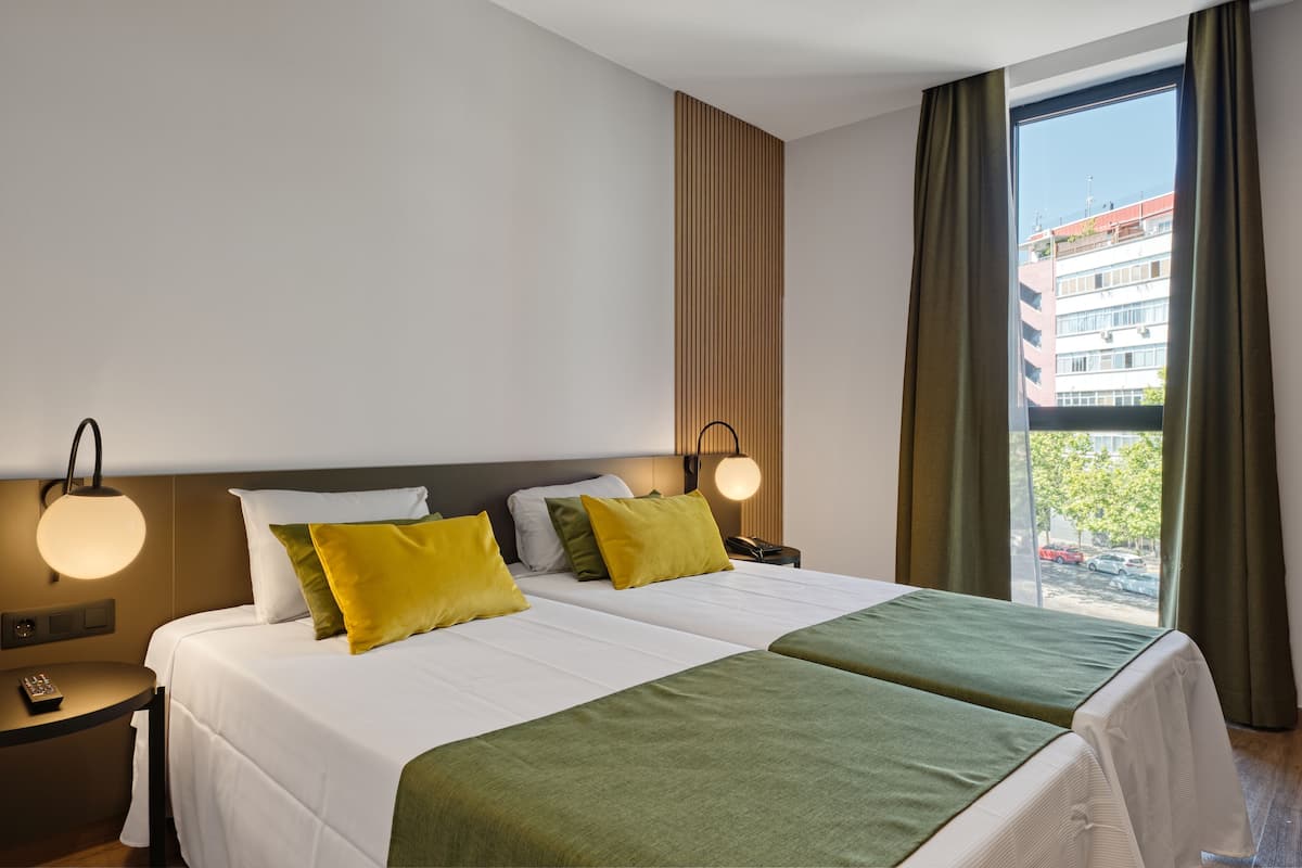 A hotel room with two beds, a nightstand, a lamp, and a view of the city through a window.