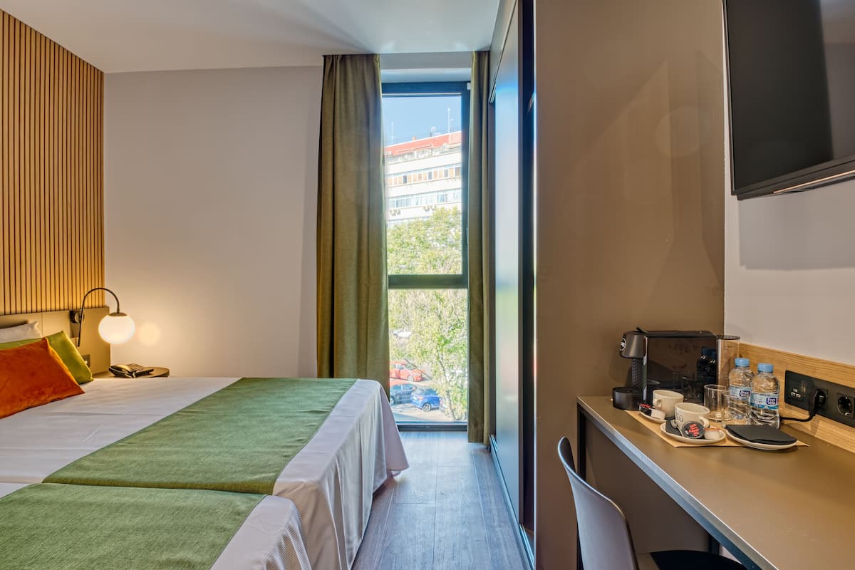A hotel room with a bed, a desk with a coffee maker, and a view of a building and trees outside.