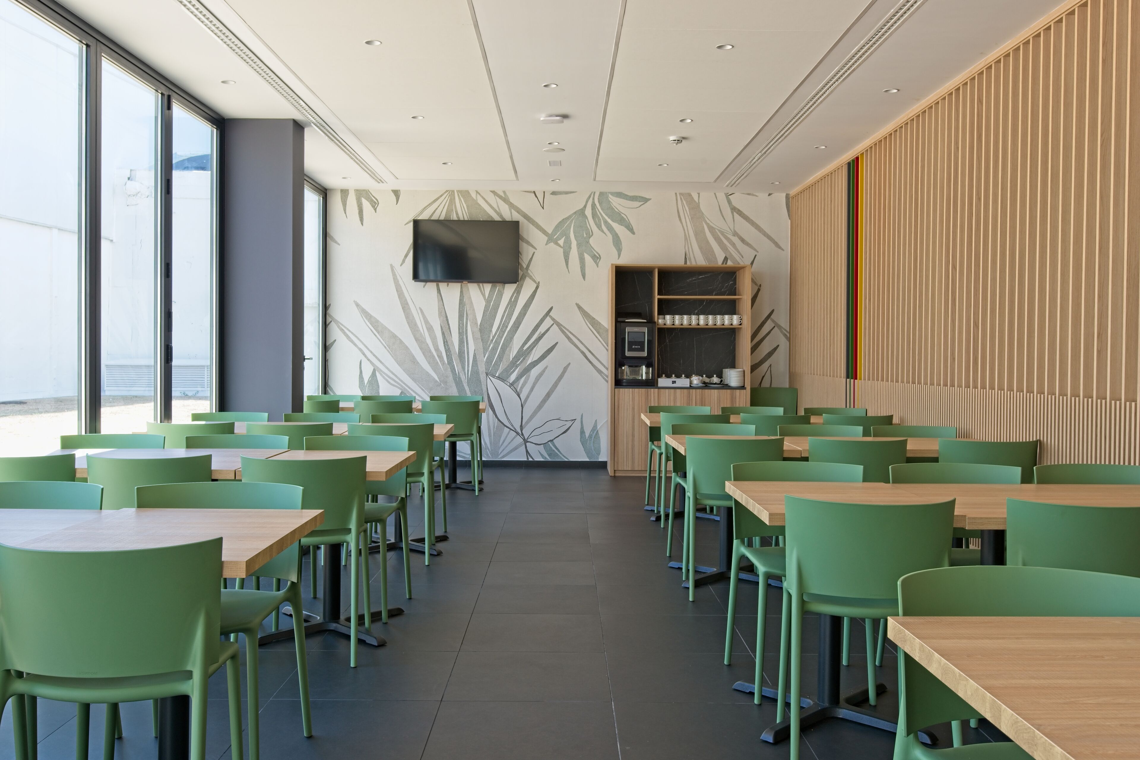 A modern dining area with green chairs and wooden tables, a flat-screen TV mounted on the wall, and a decorative wall with leaf patterns.