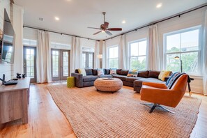 A spacious living room with a large sectional sofa, a round ottoman, and a modern coffee table. There is a flat-screen TV mounted on the wall, a desk with a lamp, and a ceiling fan.