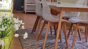 Ruokailutilassa on puinen pöytä ja valkoiset tuolit, värikäs matto ja seinällä taulu.