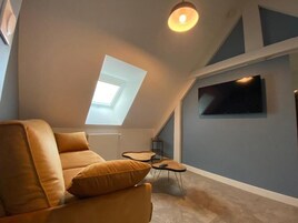 Un dormitorio tipo loft moderno con techo inclinado, tragaluces, una cama grande, televisor montado en la pared y una pequeña mesa con una silla.