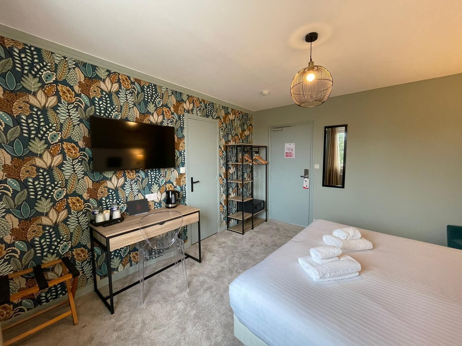 A hotel room with a floral wallpaper, a bed with white towels, a desk with a coffee maker, a TV mounted on the wall, and a hanging light fixture.