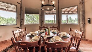 A dining area with a wooden table set for four, surrounded by wooden chairs. There are three windows providing natural light and a view of greenery outside. - White Mountain Retreat by Ruidoso Vacation Rentals (Ruidoso)