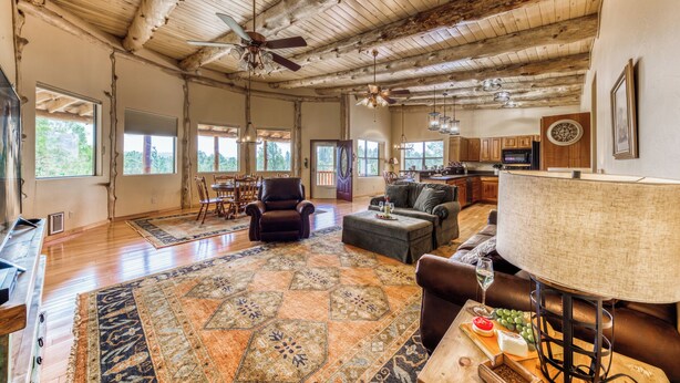 A spacious living area with a wooden ceiling, large windows, a fireplace, and a dining area with chairs and a table. - White Mountain Retreat by Ruidoso Vacation Rentals (Ruidoso)