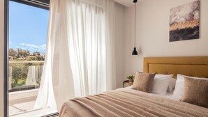 A bedroom with a large window, a bed with a beige headboard, and a painting on the wall.