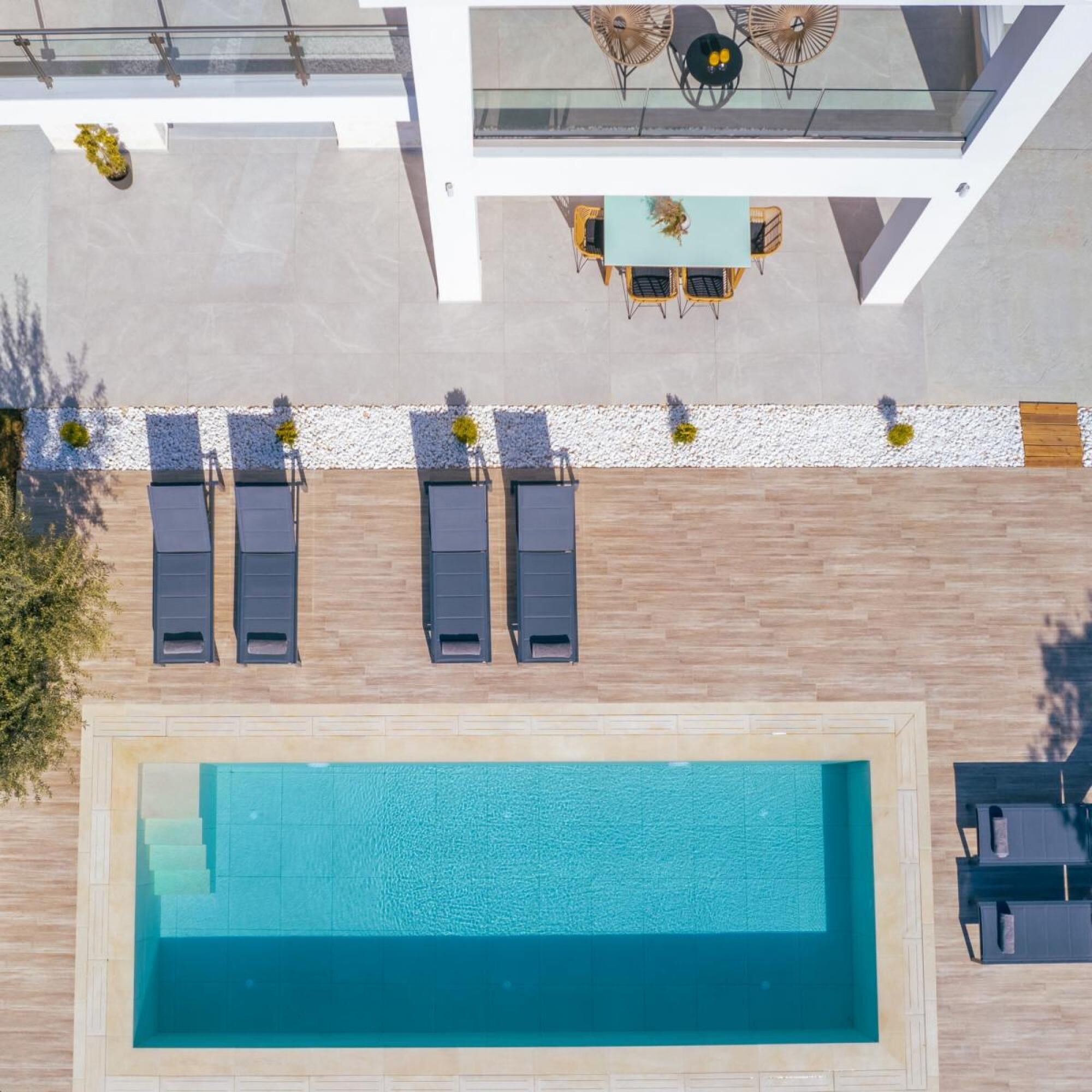 Vue aérienne d’un espace piscine moderne, avec une piscine rectangulaire, des chaises longues, une petite table avec des chaises et une rambarde blanche.