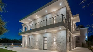 A modern two-story house with a balcony, stone exterior, and a swimming pool in the background.