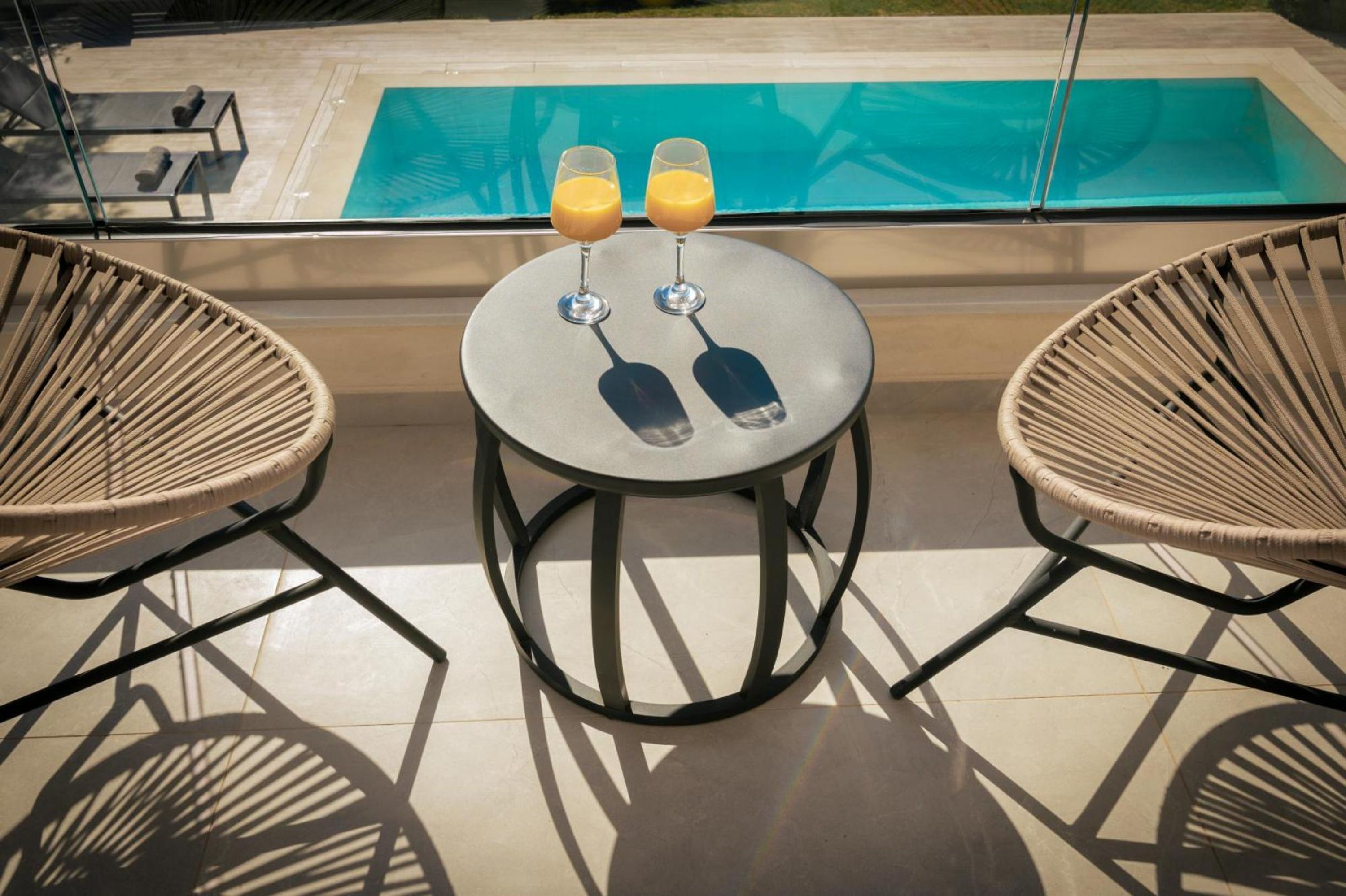 A poolside setting with two wicker chairs and a small table, each with a glass of orange juice.