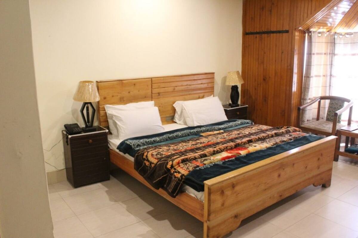 A wooden bed with a patterned blanket, two bedside tables with lamps, and a chair in a room with wooden walls and a window.