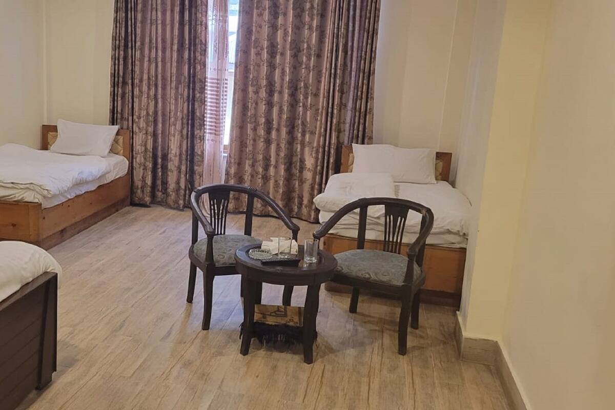A hotel room with two single beds, a small table with chairs, and a large window with curtains.
